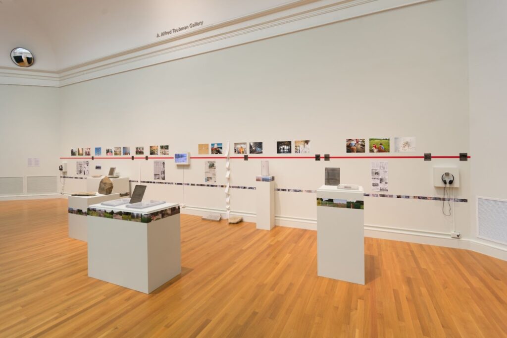 A white wall with dozens of small photographs and newspaper clippings arranged in a timeline. There are also rocks and plaques on white plinths.