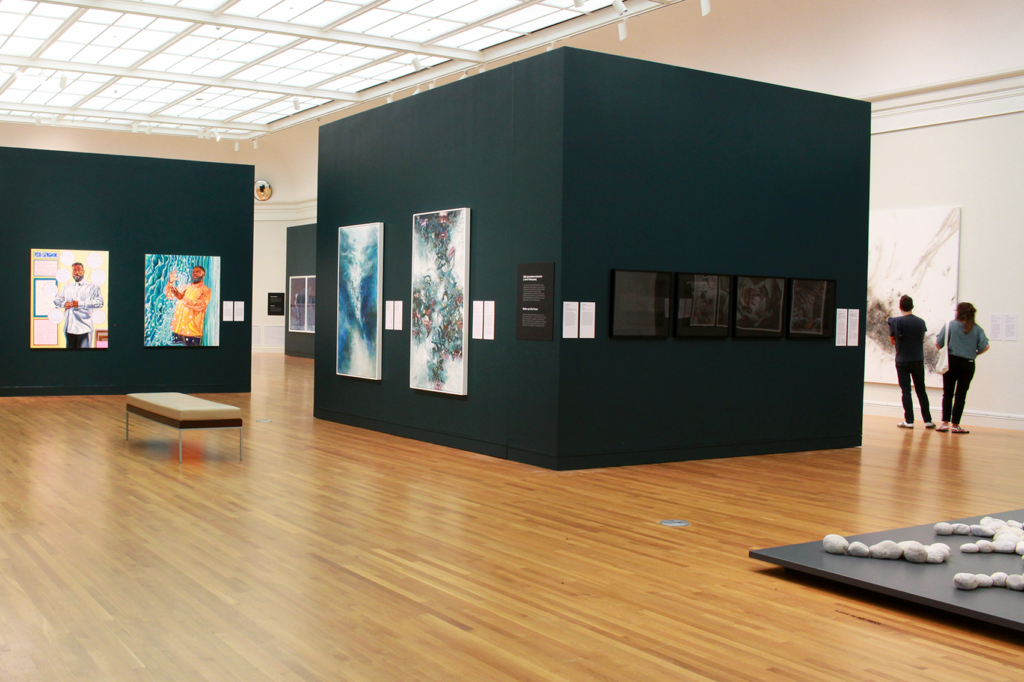 An installation view of a room with dark green walls, and several paintings. Two people stand looking at a piece.