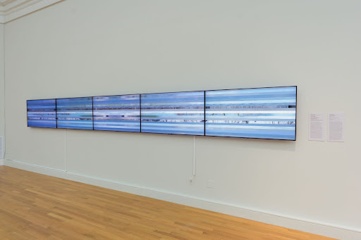 Five television screens are hanging next to each other on a white wall. The screens show strips of videos spliced together, all in shades of blue and white.