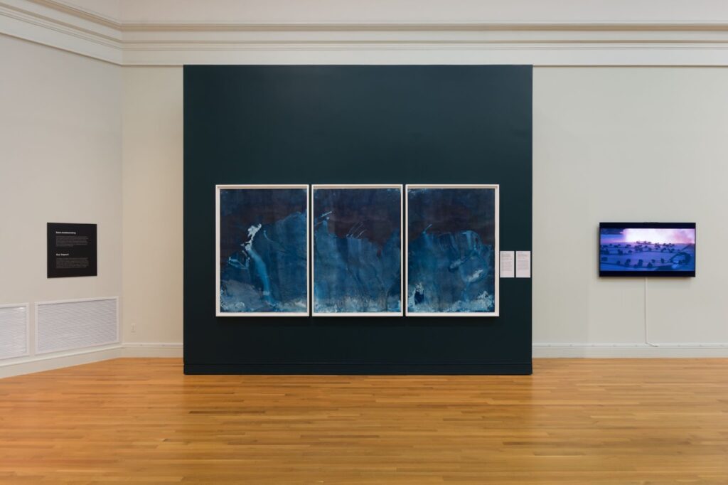 Three large framed rectangular pieces are hanging on a dark green wall. They are all shades of blue swirling together--darker on top, medium in the middle, and lighter on the bottom. To the right is a video screen which is part of a different artwork.