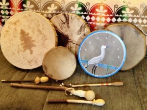 Five hand drums lined up together. One has the shape of a tree on the hide and one is painted with a crane on it. The drumsticks are in front of them.